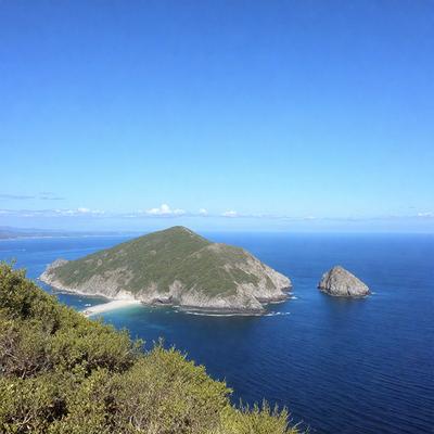 The primary subject of the image is a beautiful island in the middle of the ocean. The island is surrounded by a vast body of water, and it appears to be a lone rock in the middle of the ocean. The setting is a clear blue sky, which adds to the serene and peaceful atmosphere of the scene. The visual style of the image is a photo, capturing the natural beauty of the island and its surroundings. The colors and mood of the image are predominantly blue, with the clear sky and the